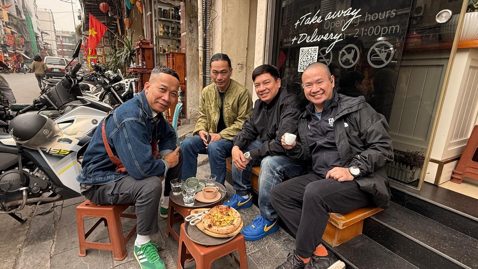 Four friends sharing coffee and pizza on a sidewalk in Hanoi’s Old Quarter
