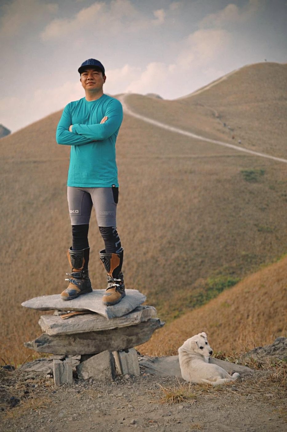 Loija in a teal shirt and riding boots standing on a stone cairn in golden hills, a white puppy resting nearby