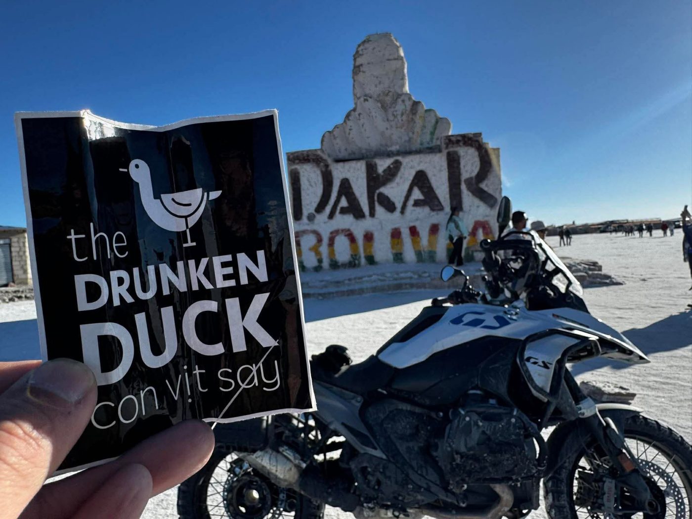 A Drunken Duck sticker held up before the Dakar Bolivia monument, with an adventure motorbike beside it