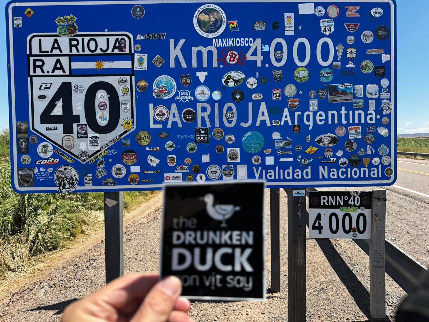 A Drunken Duck sticker held up in front of the Ruta 40 Km 4000 sign in La Rioja, Argentina